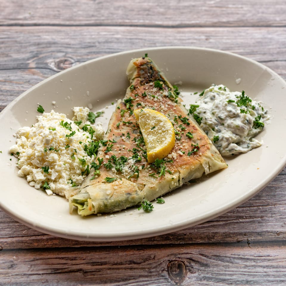 A Greek Bites spanakopita with feta cheese, tzatziki, and a lemon wedge on a white plate.