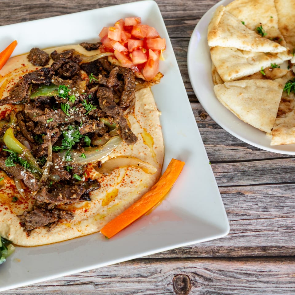 Hummus topped with beef shawarma, diced tomatoes, and pita bread on a wooden table.