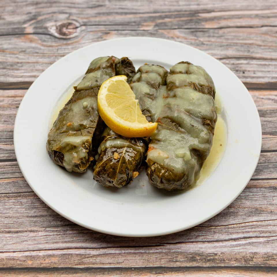 A white plate of Greek dolmades, grape leaves stuffed with rice, topped with lemon and sauce.