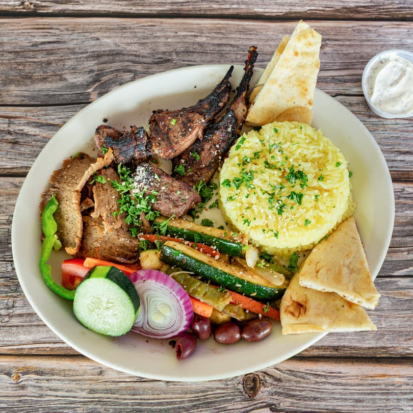 A Greek Bites platter with grilled lamb chops, gyro meat, rice, pita, vegetables, and olives.